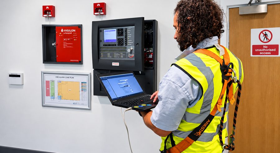 Engineer testing a Honeywell fire alarm panel with a laptop, beside the fire alarm zone plan
